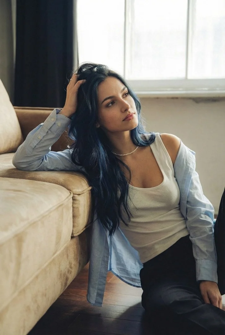 A young woman with long dark hair with blue highlights, wearing a white tank top, a light blue shirt, and a pearl necklace, sitting on the floor next to a beige sofa in front of a window, looking thoughtful with her hand resting on her head.
