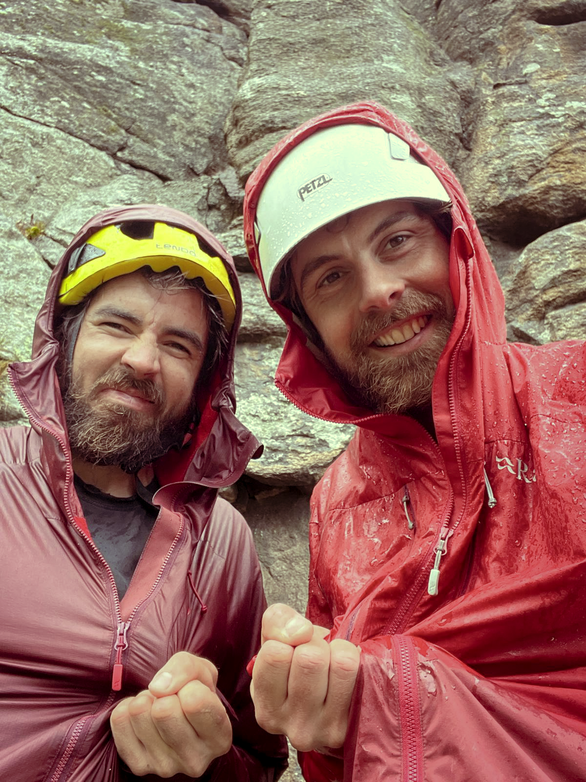 Two men in rain jackets and helmets smiling and shaking hands in front of a rocky background.