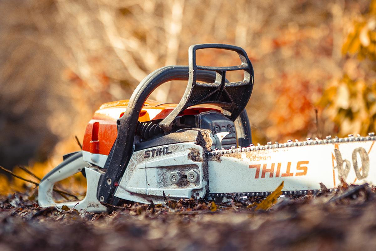 A chainsaw resting on fallen leaves in an outdoor setting with blurred autumn foliage in the background.