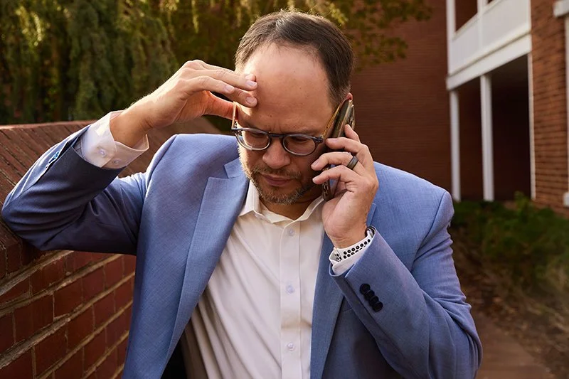 A man in a blue suit and glasses is talking on a cell phone while holding his forehead with a worried expression.