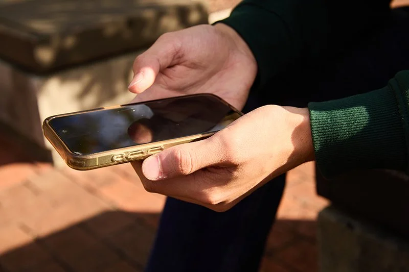 Close-up of person holding a smartphone with a golden case, sitting outdoors in sunlight.