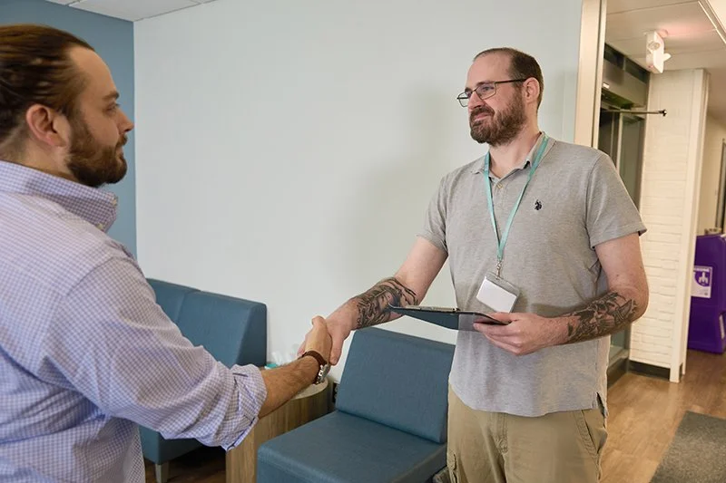 Two men shake hands in a professional setting, one holding a clipboard, with a waiting area in the background.