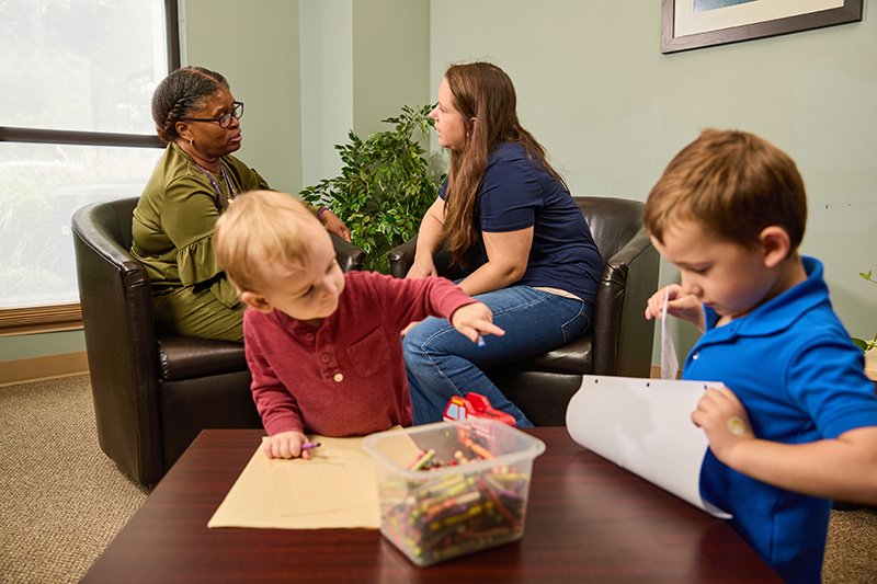 A therapist and a woman are having a conversation while two children are drawing at a table with crayons at the Maryland Health Association