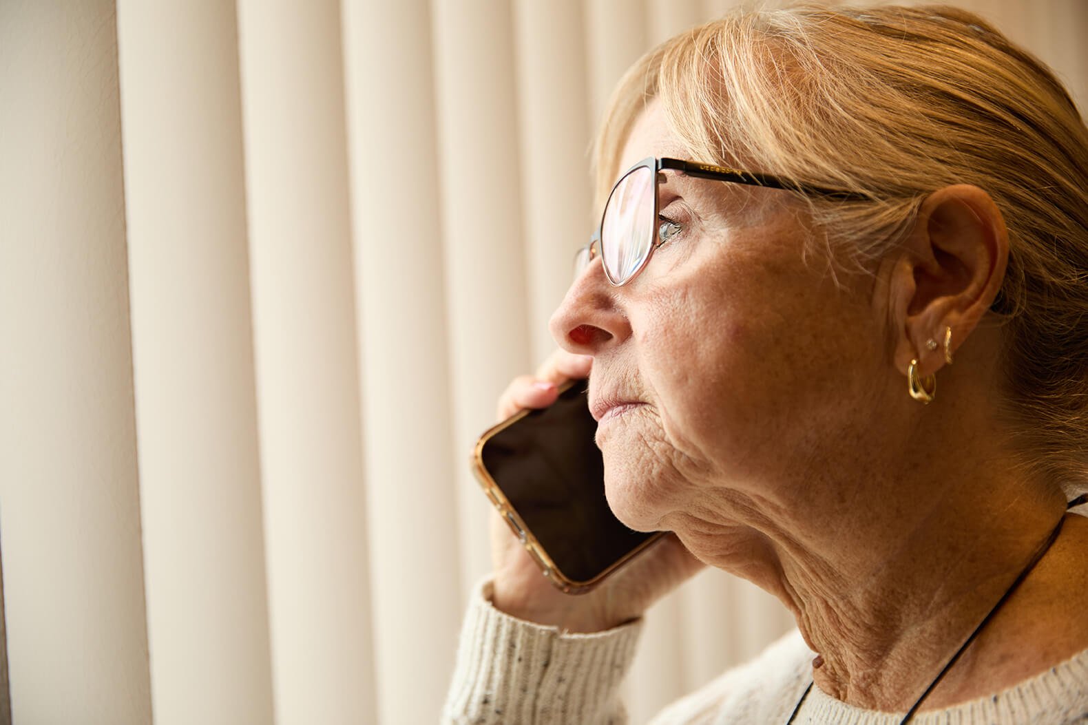 An elderly woman with glasses talking on the phone, looking out a window with vertical blinds.