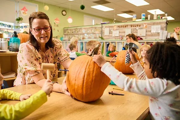 Woman in cheerful classroom helps children decorate a pumpkin