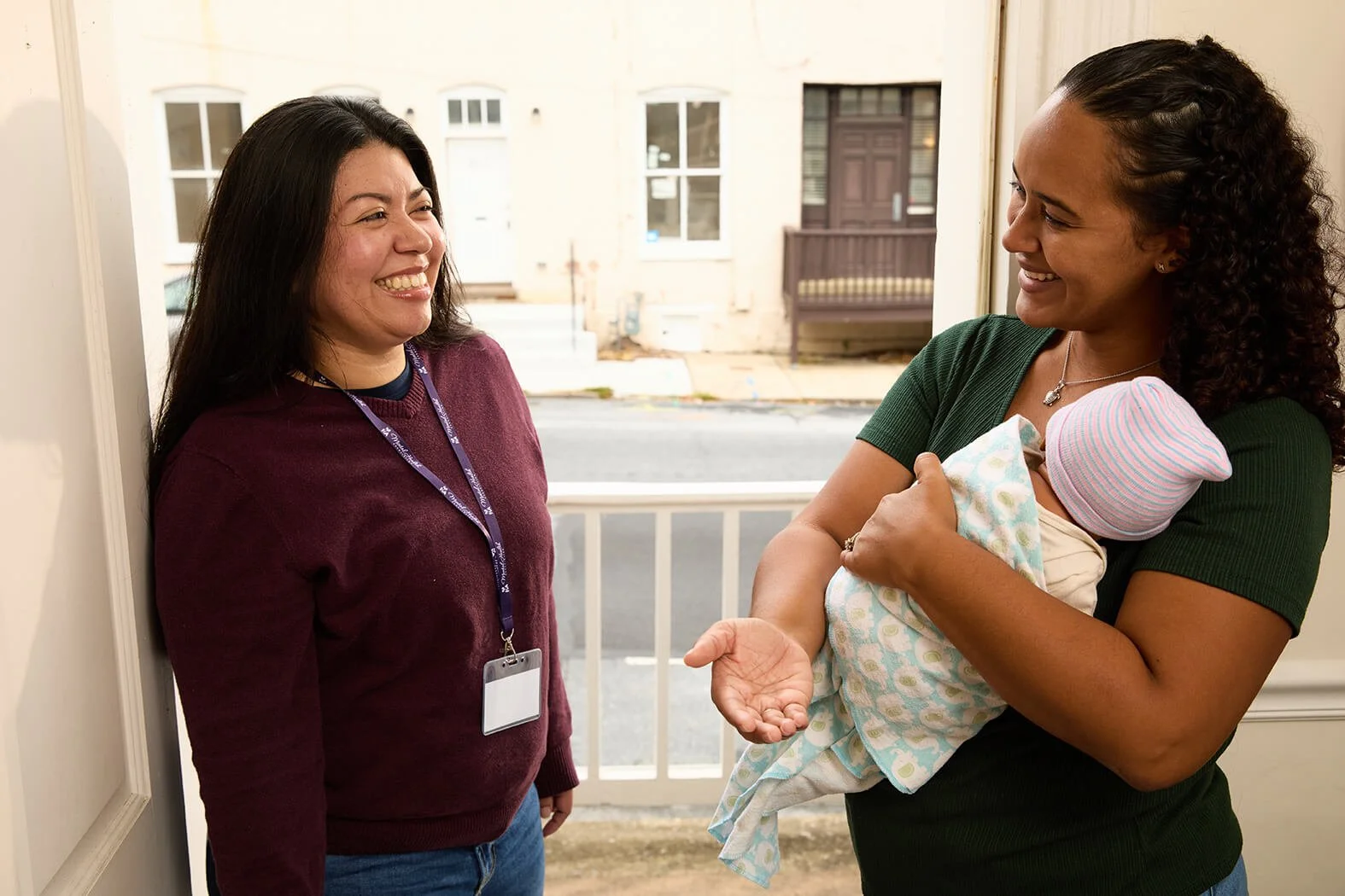 Two women standing by a door, one holding a baby, having a conversation and smiling.