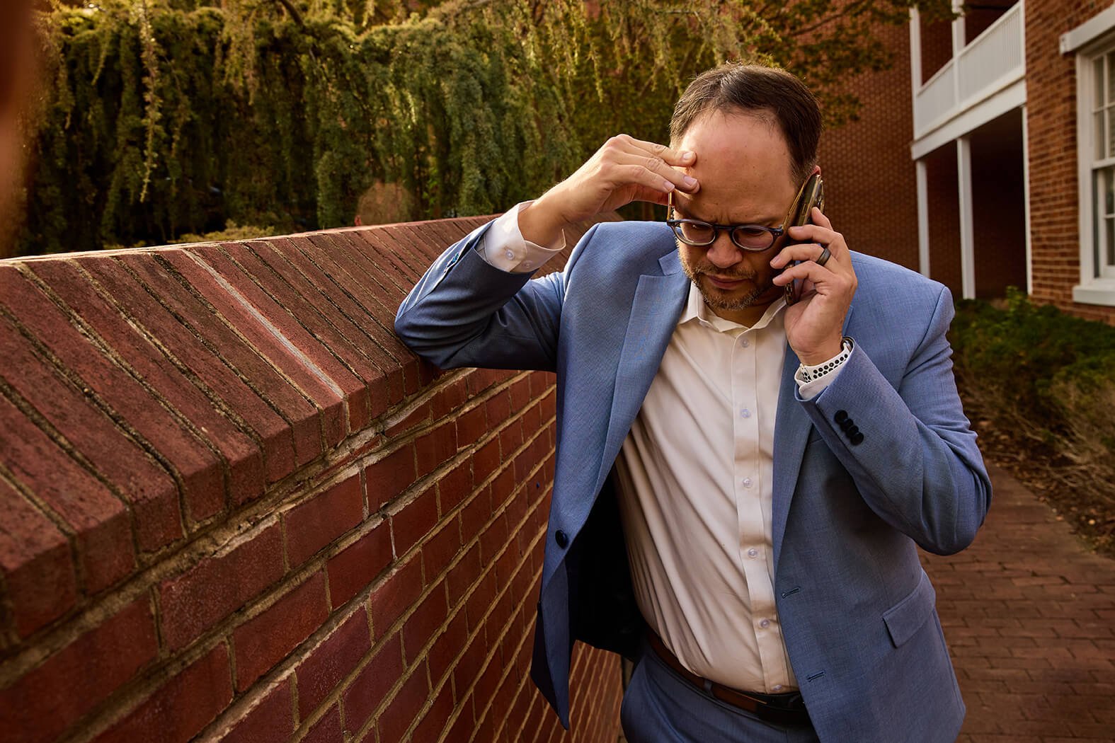 A man in a blue suit and white shirt leaning against a brick wall, holding a phone to his ear, appearing stressed