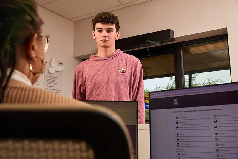 A young man in a pink hoodie standing in the reception area of the Frederick County MHA, facing a woman sitting at a desk with computer screens in front of her.