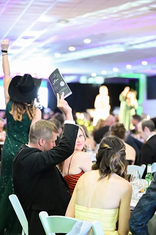 People at the Catoctin Affair. One man raising his hand and a woman in a green dress with a black hat dancing in the background. Other guests sit at tables with drinks 