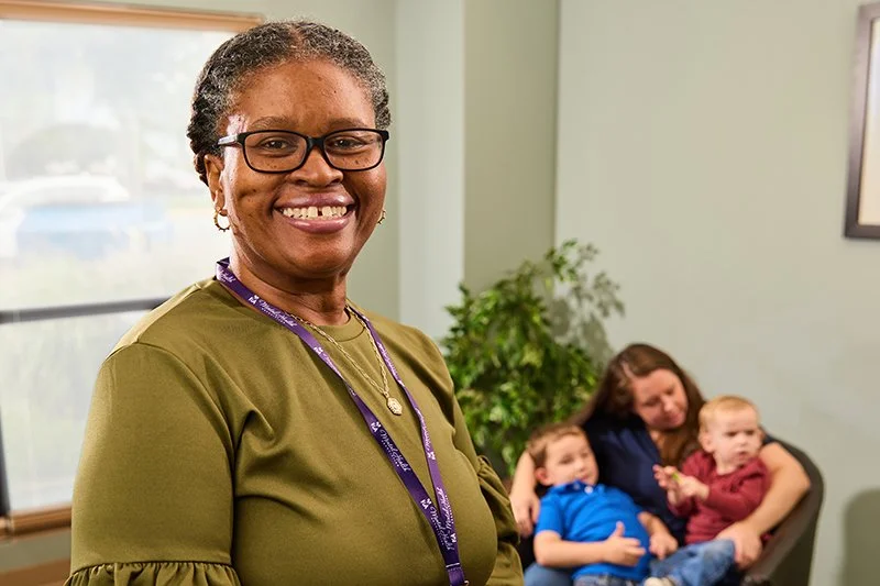 smiling woman with glasses and a green shirt standing in a room with a window, a plant, and a woman sitting with two children in the background.