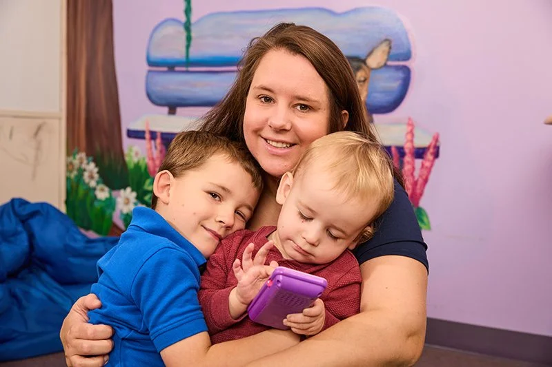 A woman with long brown hair hugging two young boys in a fun looking therapist's office
