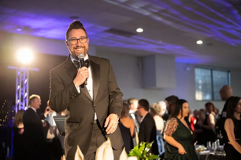 A man in a tuxedo with glasses holding a microphone and smiling at  the Catoctin Affair with other well-dressed guests and colorful lighting.