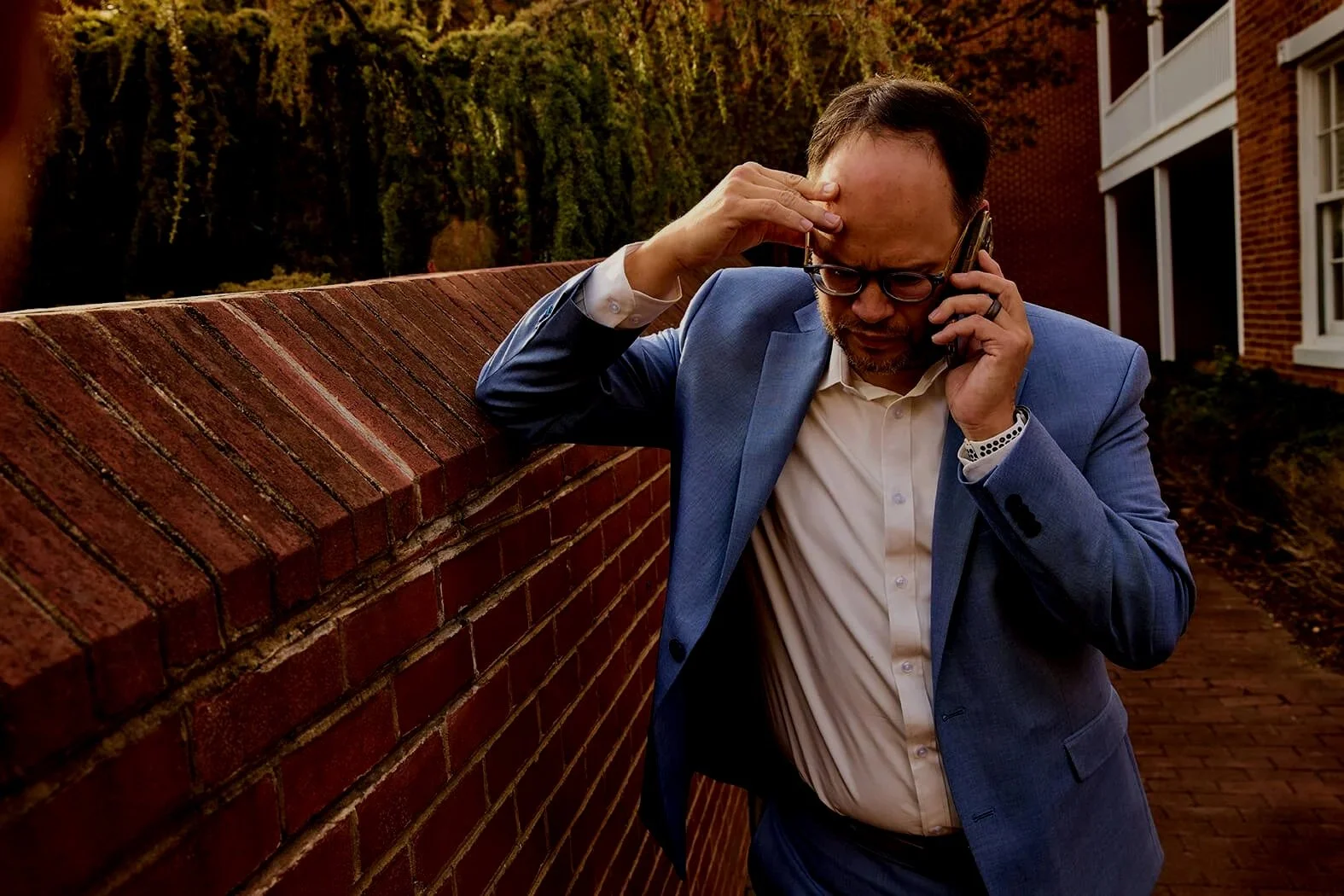 A man in a blue suit and white shirt leaning against a brick wall, holding a phone to his ear, appearing stressed