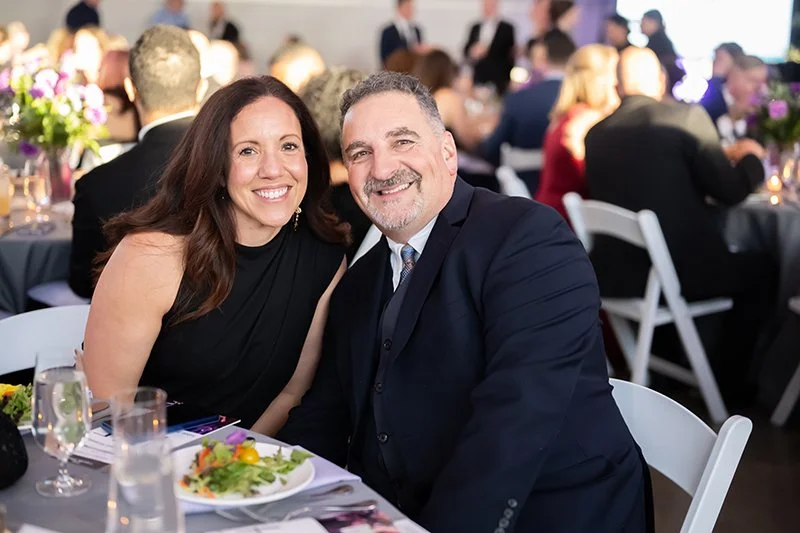 A smiling couple sitting at a banquet table during  the Catoctin Affair with other guests in the background. 