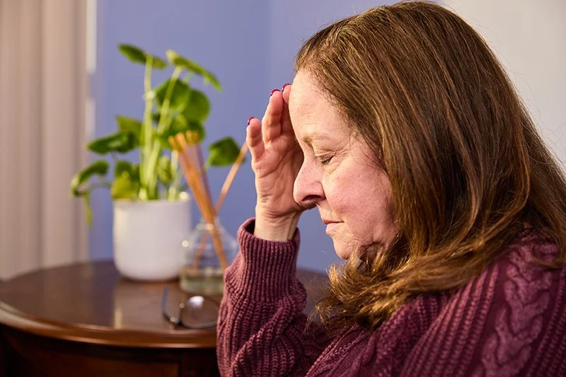 A woman with brown hair and a maroon sweater sits with her eyes closed, holding her head with her right hand, appearing distressed.