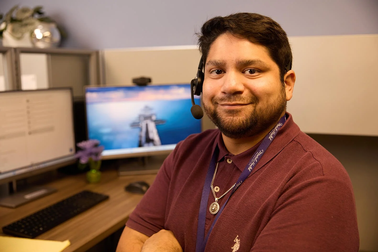 A man with a headset smiling in an office cubicle, with dual monitors and a keyboard on the desk.