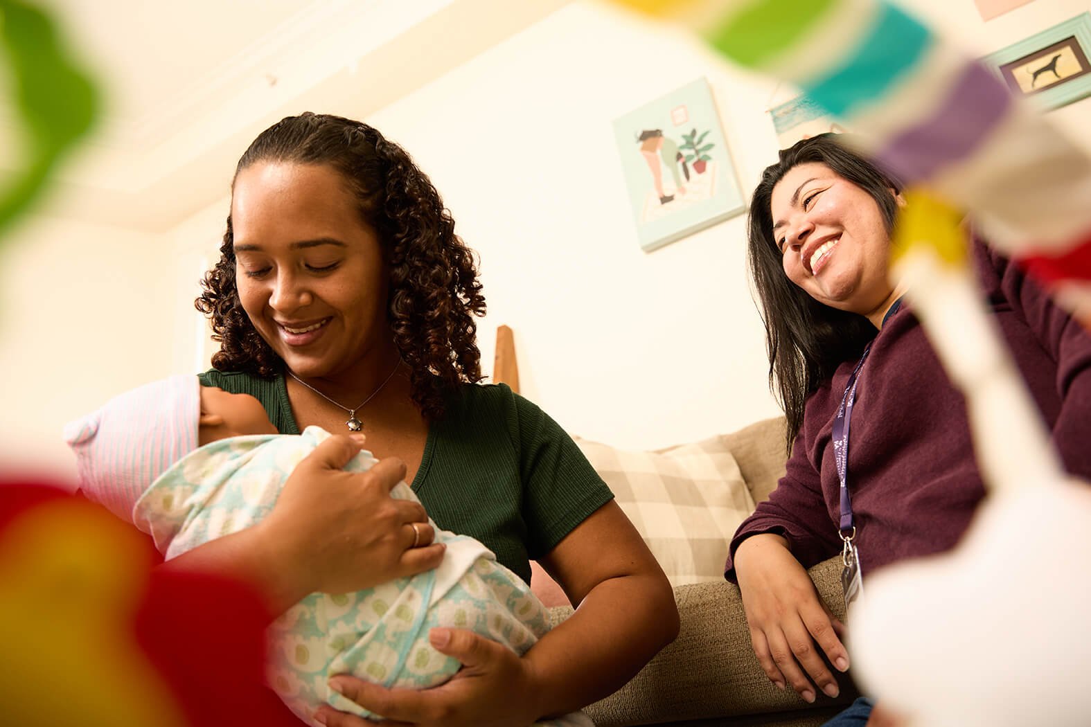 A woman holds a newborn baby wrapped in a blanket, with another woman sitting nearby smiling.