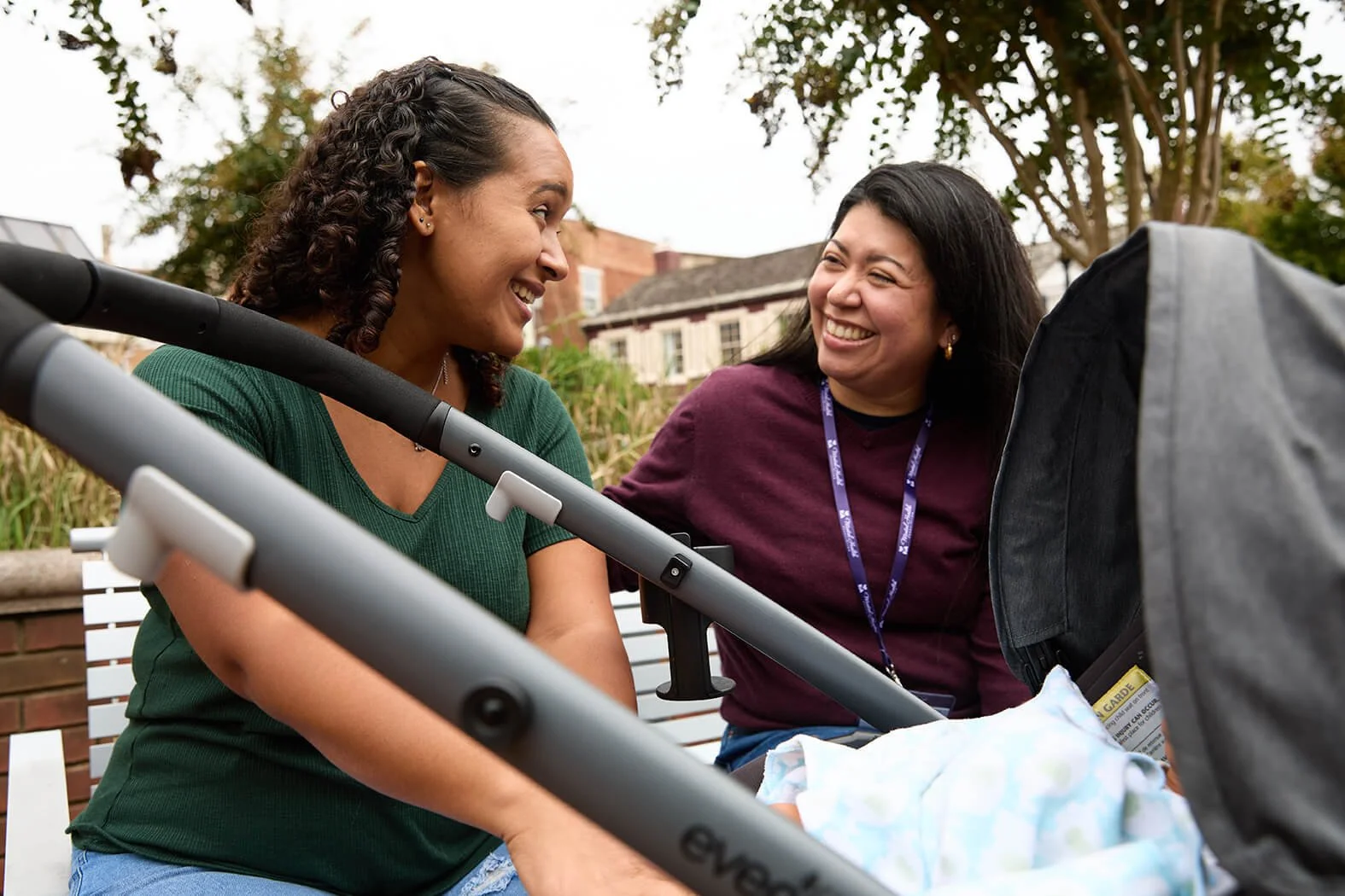 Two women smiling and talking to each other outdoors, with a baby stroller in front of them and a blanket inside. One woman has a lanyard from the Maryland Health Associates to signify that she is a counselor there
