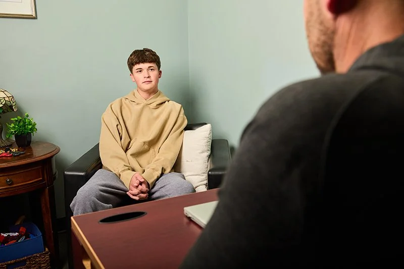 A young boy sitting on a black sofa with a white pillow, during a therapy session at the Mental Health Association