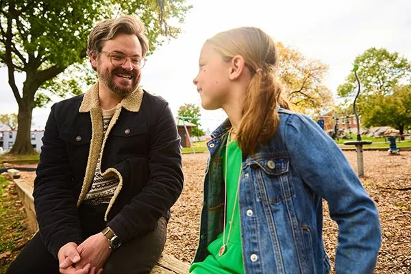 Smiling man with beard and glasses sits outside and talks to young girl with blond pigtails