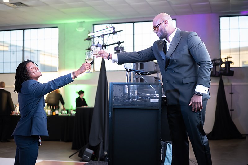 Two people in formal attire, a woman and a man, standing on a dance floor and clinking glasses. 