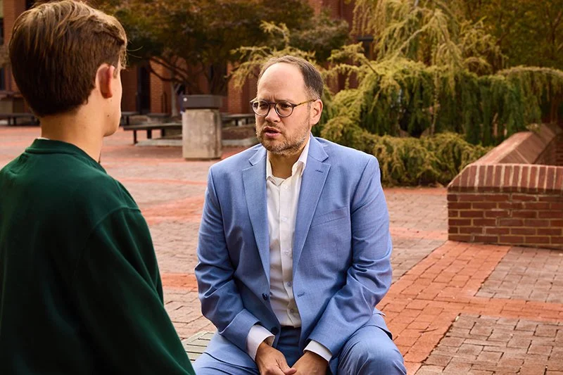 A man in a light blue suit and glasses talking to a young person with brown hair wearing a dark green jacket outdoors on a brick plaza with trees and benches in the background.