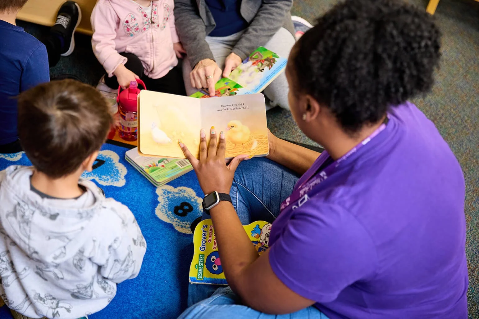 A group of children and an adult reading a children's picture book about ducks, sitting on a blue rug with a colorful pattern, in a classroom or library setting.