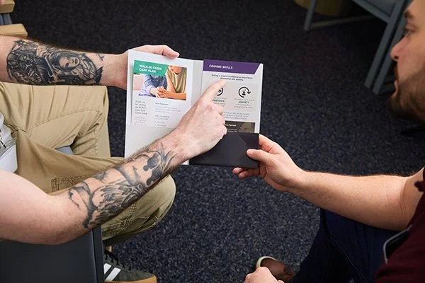 tattooed arms hold a brochure and point to it for another person