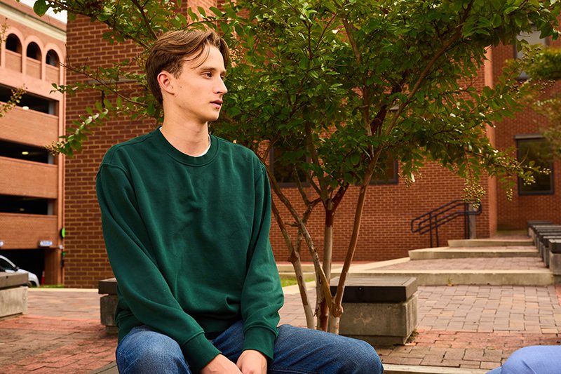 A young man with short light brown hair, wearing a green sweatshirt and blue jeans, sitting outdoors in thought