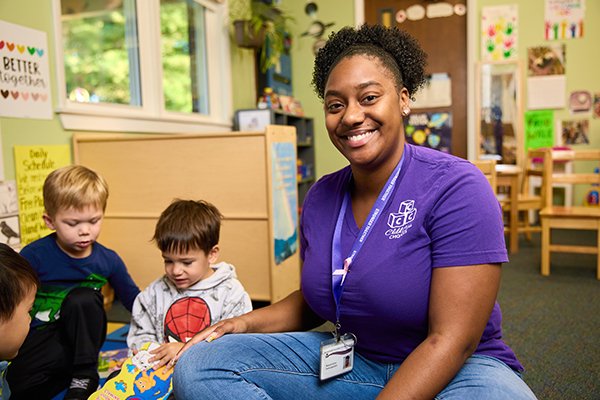 Woman sits on floor with young children in a classroom while wearing t shirt Children Care Choices