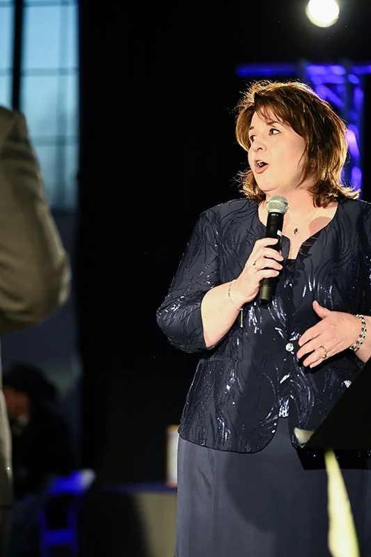 A woman with shoulder-length brown hair holding a microphone and speaking at the Catoctin Affair