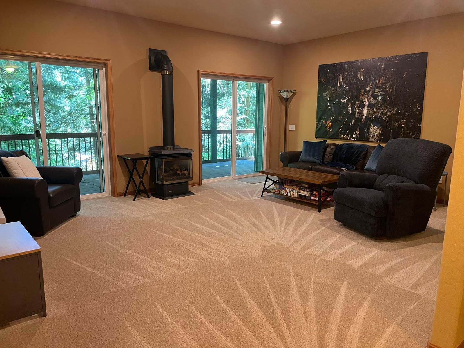 Living room with beige carpet, black leather armchair, dark blue sofa, wooden coffee table with books, and a large aerial cityscape photograph on the wall. Two sliding glass doors lead to a balcony with trees outside, and a black wood stove with a pi