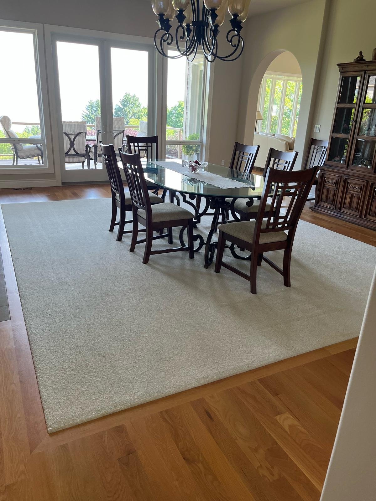 Dining room with a glass-top table, eight wooden chairs, a chandelier hanging above, a large rug, and doors leading to a balcony with outdoor chairs.