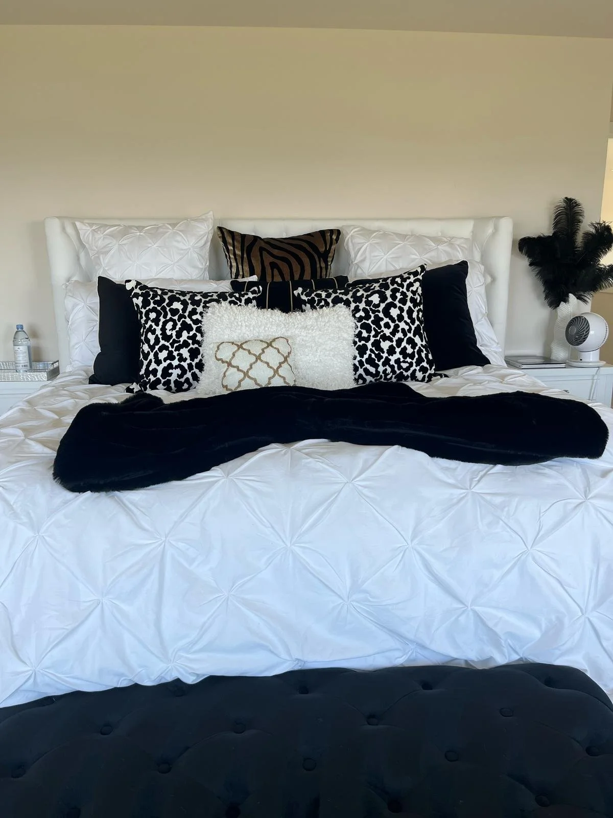 A neatly made bed with a white tufted headboard, decorated with black and white patterned pillows, and a black throw blanket at the foot. There are nightstands on either side, each with a lamp and decorative items, and a beige wall behind the bed.