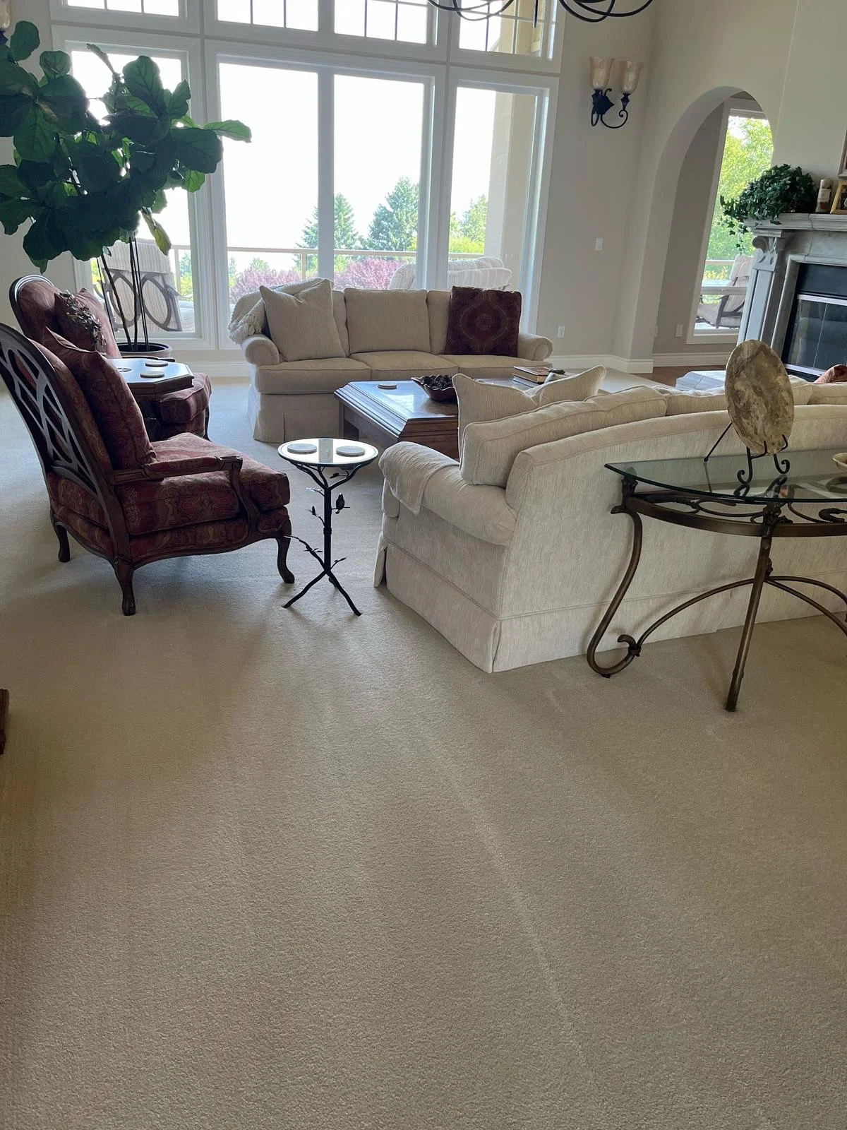 Living room with beige carpet, white couches, red armchair, wooden coffee table, glass side table, large windows showing trees, fireplace, and decorative items.