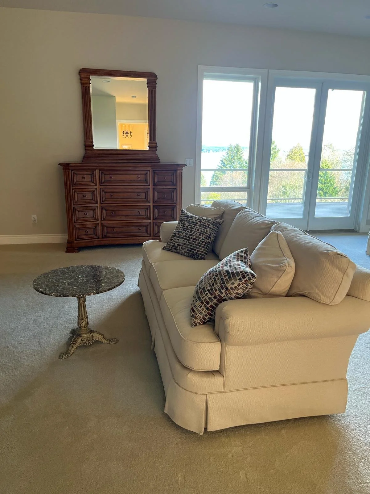 Living room with a beige sofa with patterned pillows, a small round side table with a marble top, a wooden dresser with a mirror, and large windows and glass doors showing a view of outdoor trees and water.