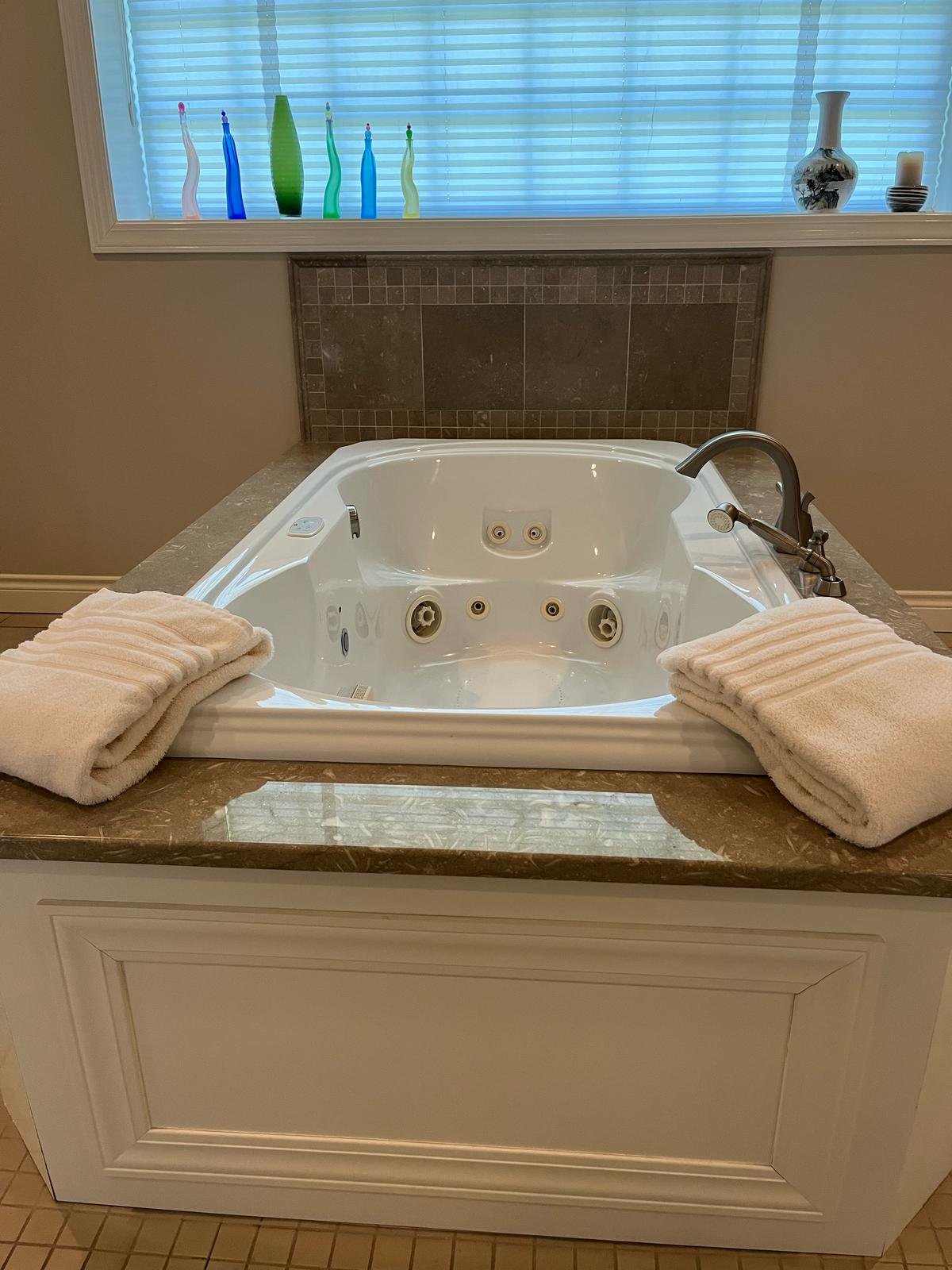 A white bathtub with built-in jets and a faucet, covered with two folded white towels, set in a bathroom with a window above it decorated with colorful glass bottles and vases.