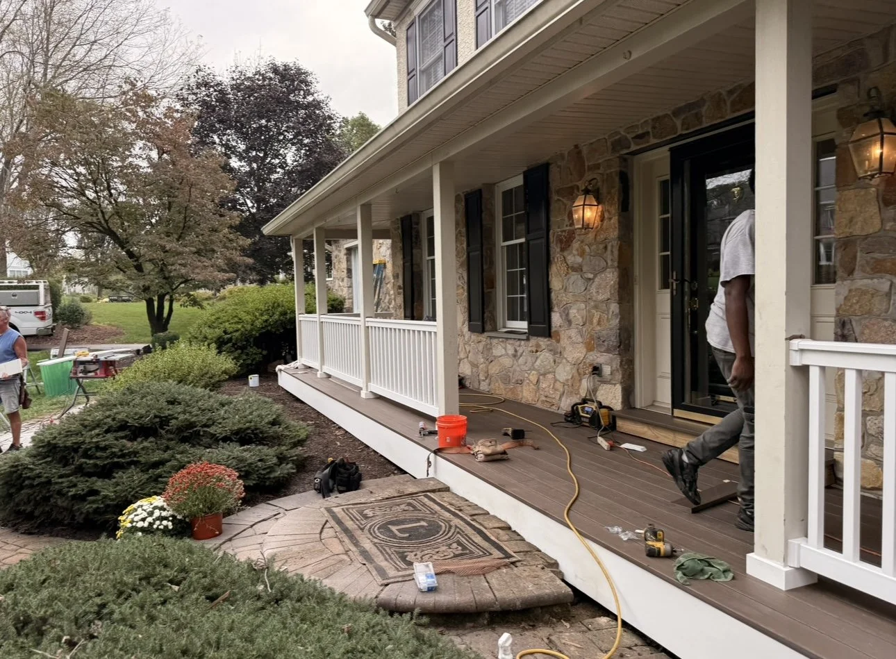Workers installing or completing a porch on a house with a stone exterior, black shutters, and outdoor lighting. Construction tools and equipment are visible on the porch and yard.