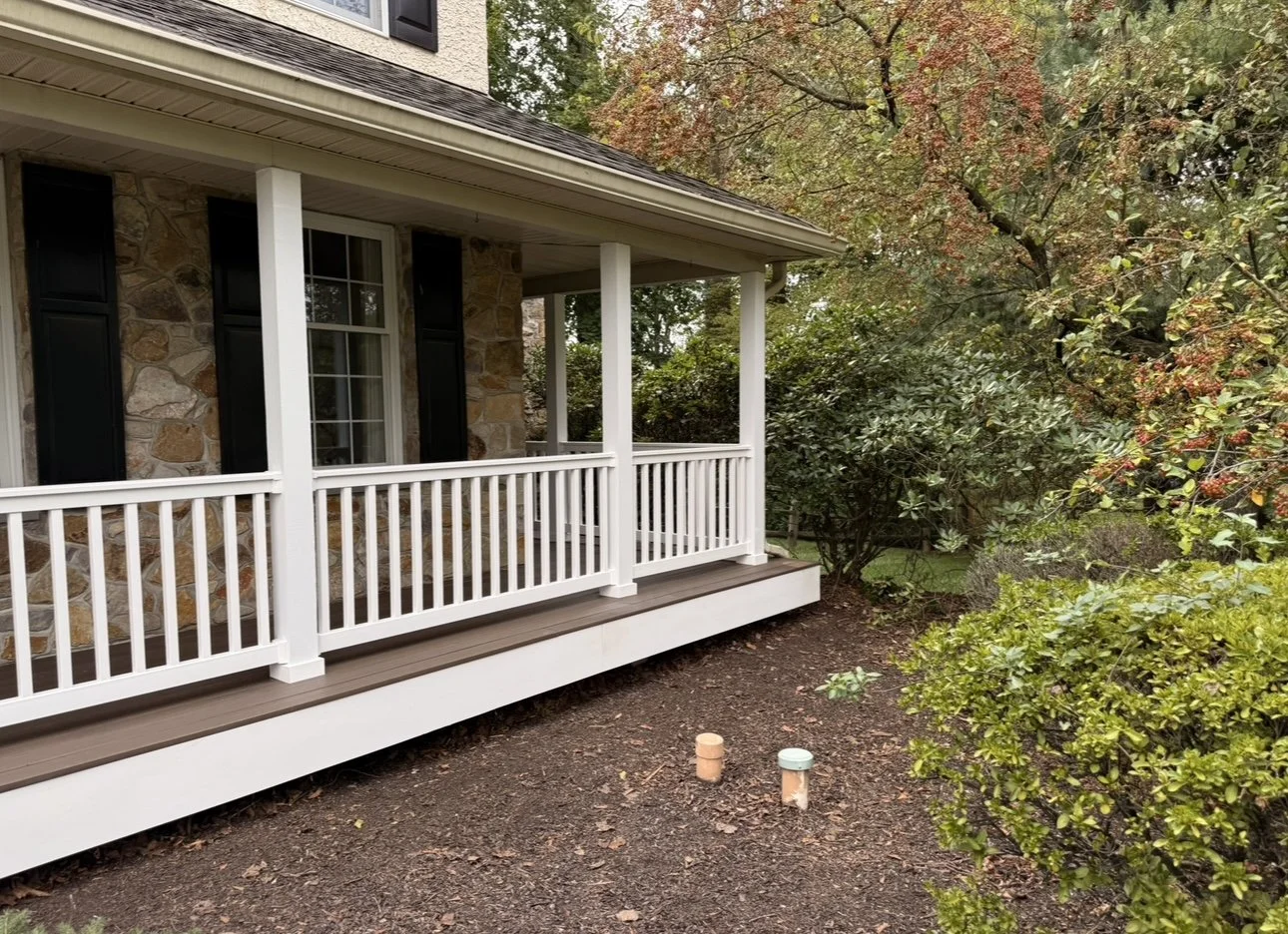 A house with a stone exterior, black shutters, white railing porch, surrounded by trees and bushes, in an autumn setting.