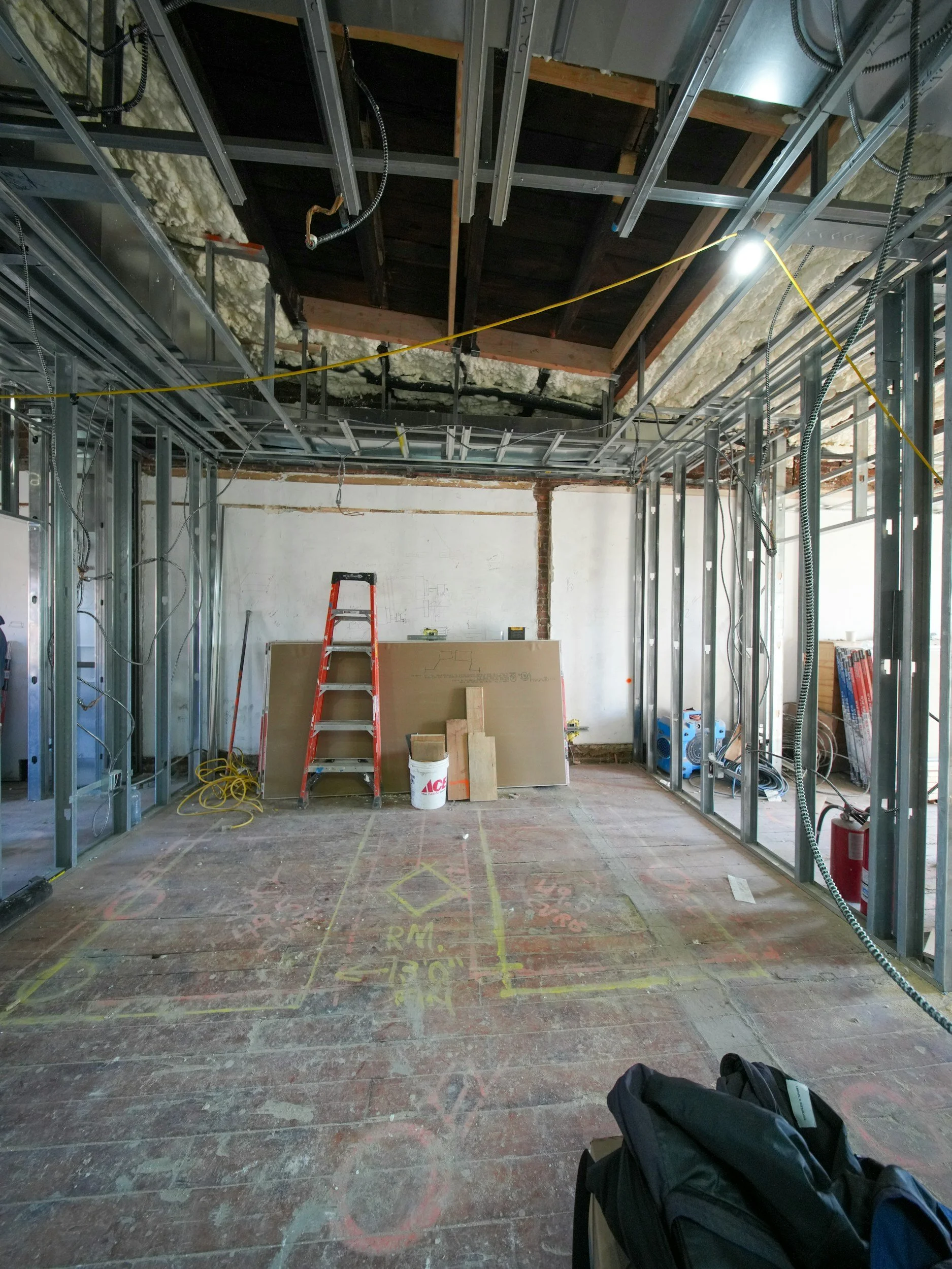 Interior view of a construction site showing unfinished walls and ceiling with exposed metal framing, pipes, and insulation. Construction tools, a ladder, and building materials are present on the floor.