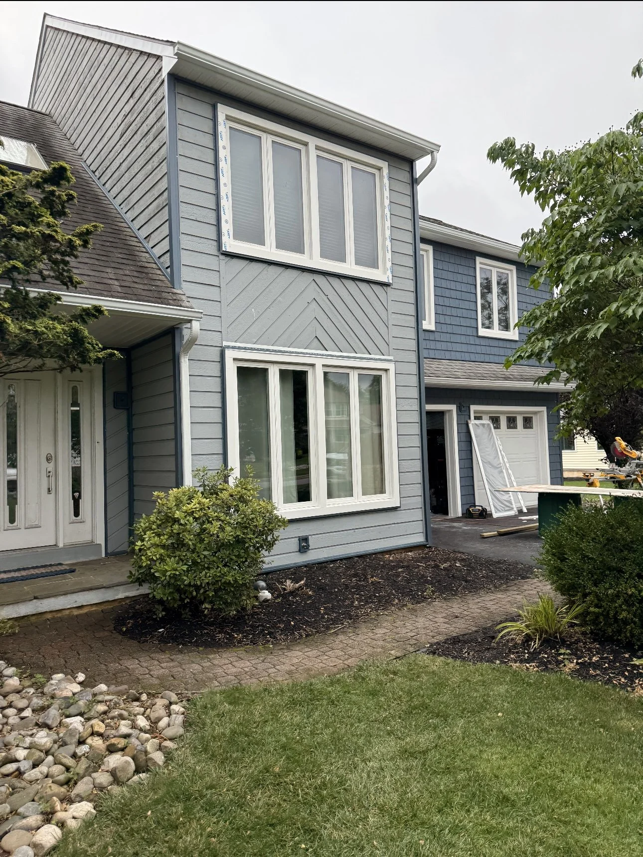A two-story house with gray siding, white window frames, and a small front yard with bushes, a pathway, and a lawn. Part of the house's exterior appears to be under construction or renovation.