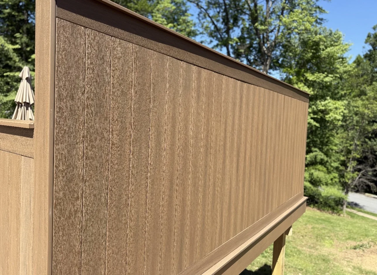 Close-up of a wooden deck railing with vertical wood paneling, against a backdrop of trees and blue sky.