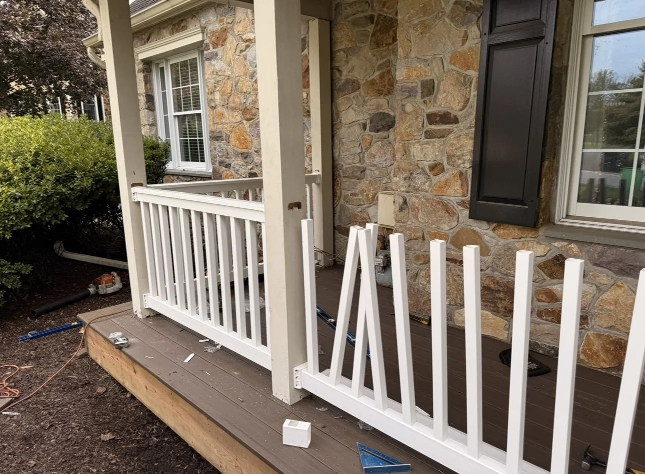 Construction of a small porch with a white railing in front of a house with a stone exterior, black shutters, and a window.