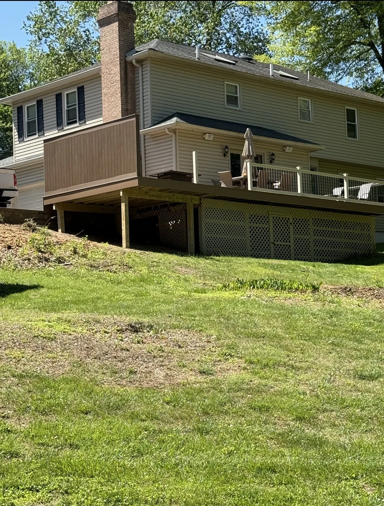 Photo of a house with a raised wooden deck, outdoor furniture, a patio umbrella, and a grassy yard in a suburban neighborhood.