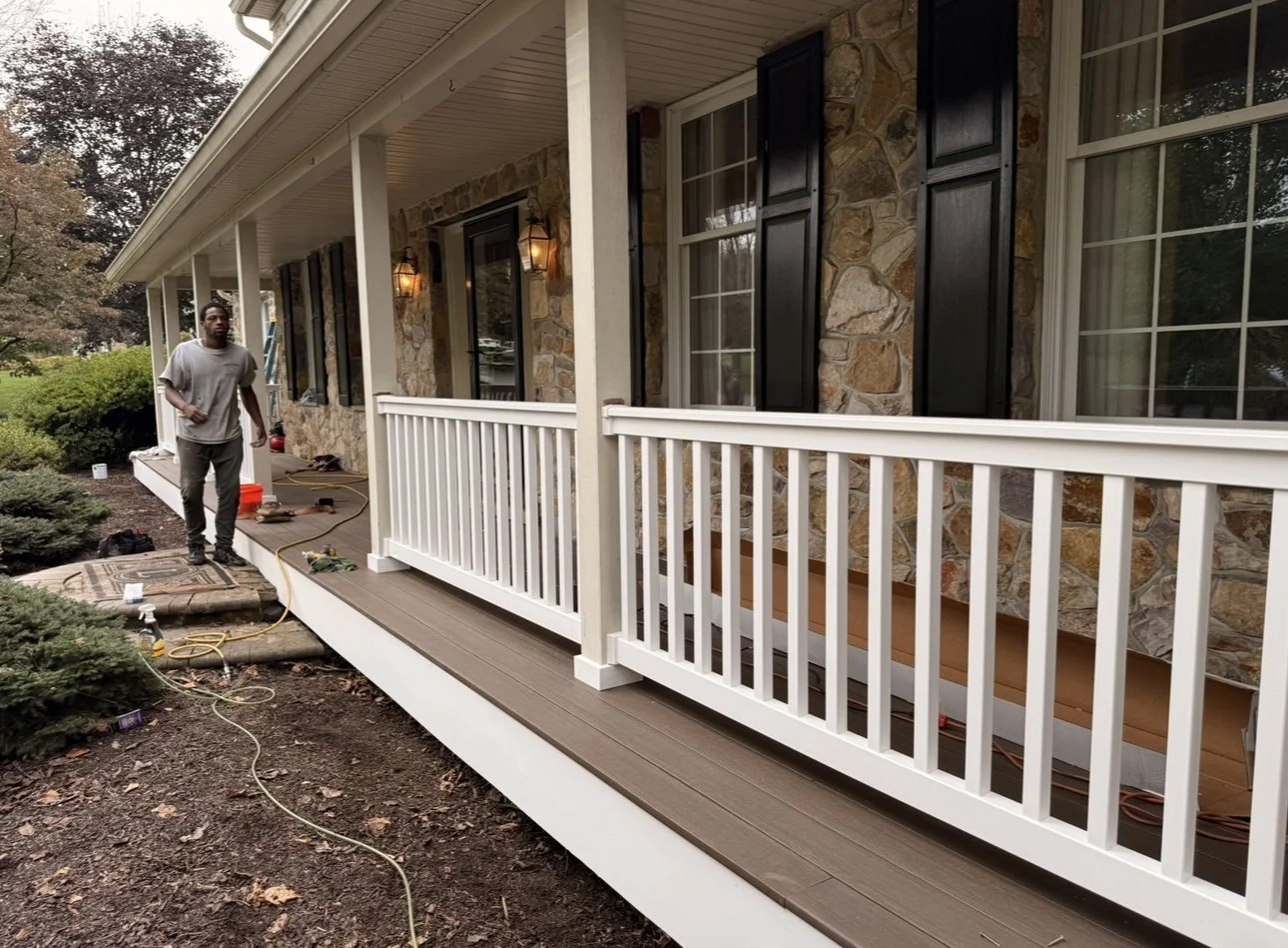 A man standing on a porch under construction of a house, with tools and equipment around, in front of a stone house with black shutters and a white railing.