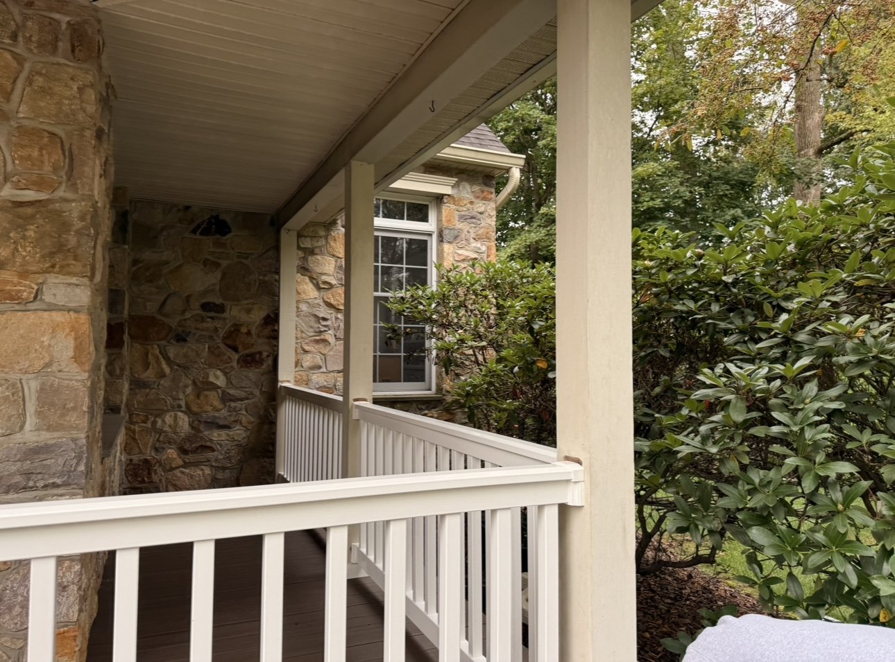 A porch with a white railing and a stone wall. Green bushes and trees are visible, with a window on the house's exterior wall.