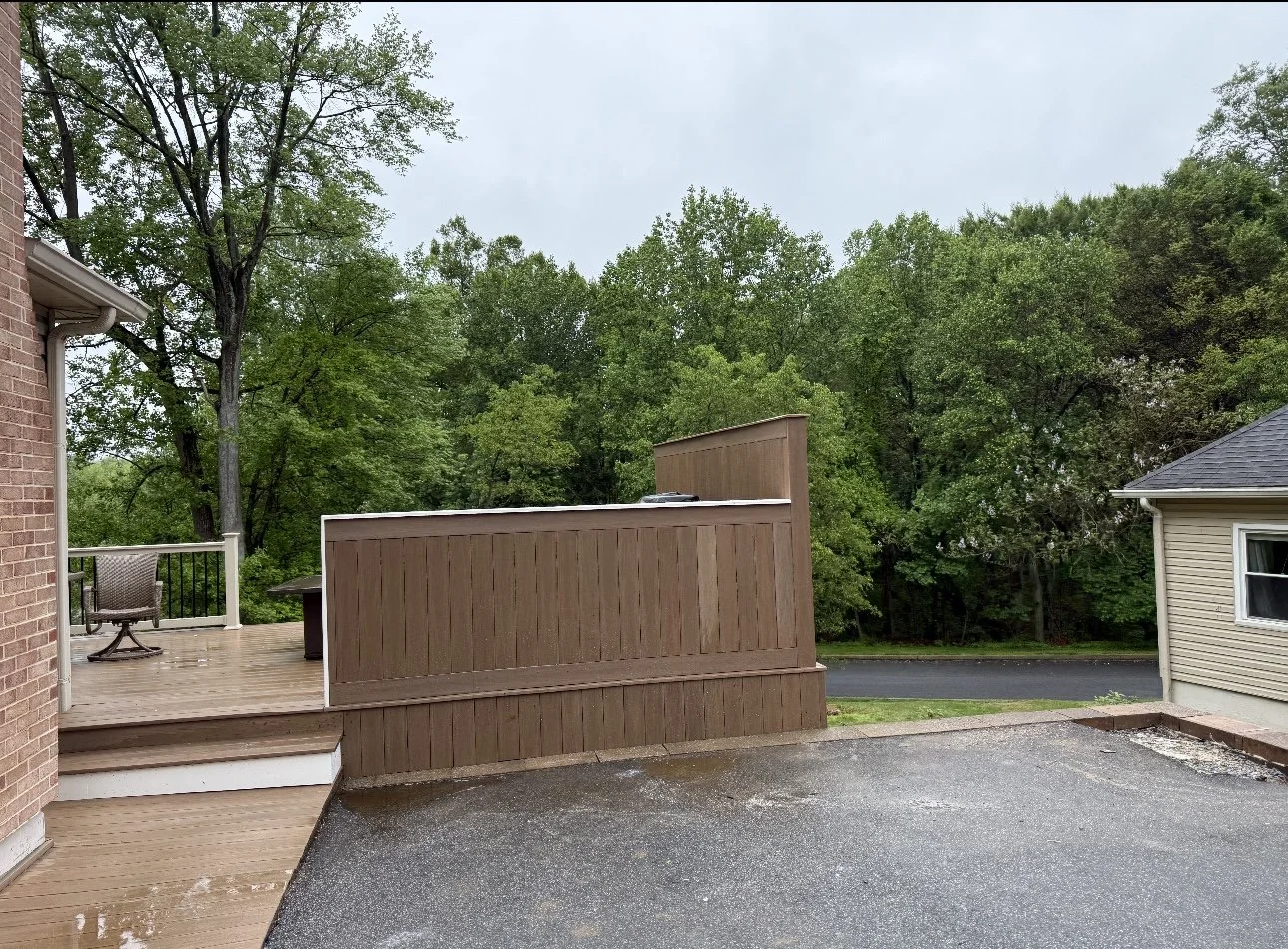 A backyard patio with a wooden deck and a brown privacy fence. The deck has chairs and a small table, with a wet surface indicating recent rain. There's a paved driveway next to the deck, and a lush, green wooded area in the background. Part of a bri
