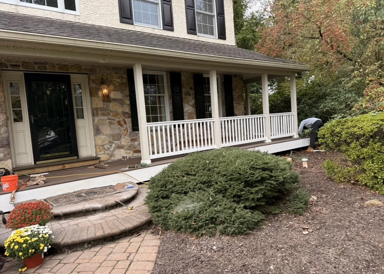 Person working on a house's front porch, which has white railings and a stone exterior, surrounded by trees with autumn-colored leaves, and decorative plants near the steps.