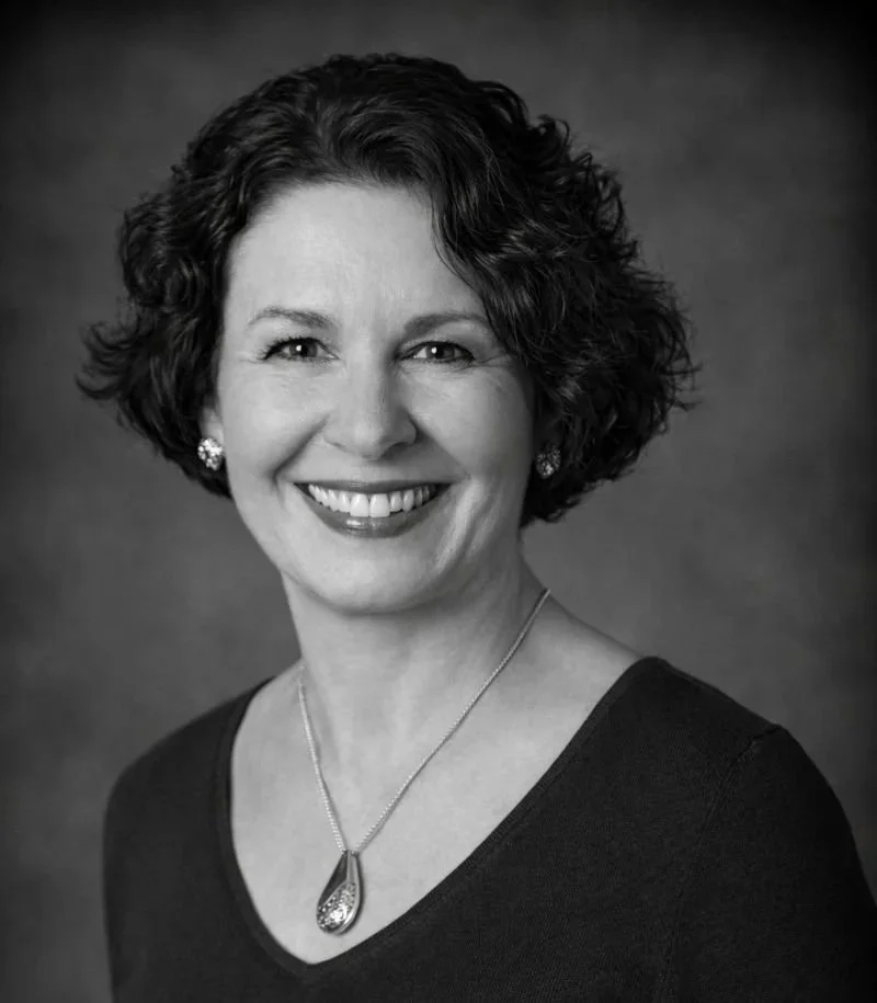 Black and white portrait of a smiling woman with short, curly hair, wearing earrings and a necklace, against a plain background.