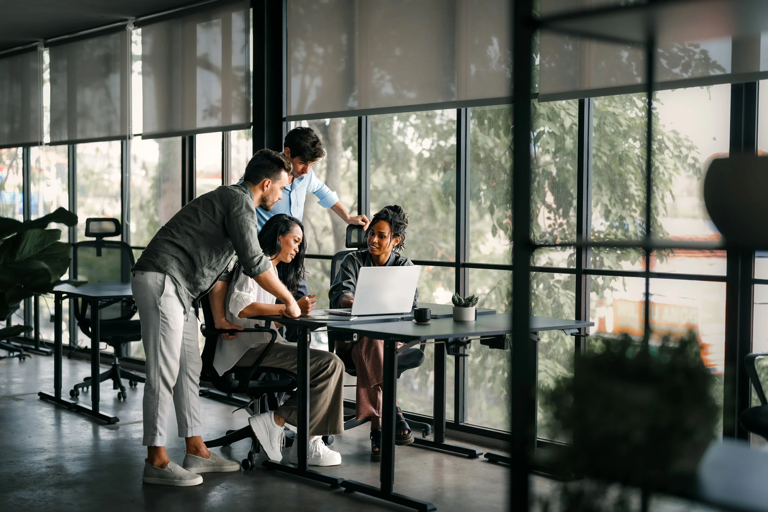 Four colleagues collaborating around a laptop and smartphone in a modern office with large windows and natural light.